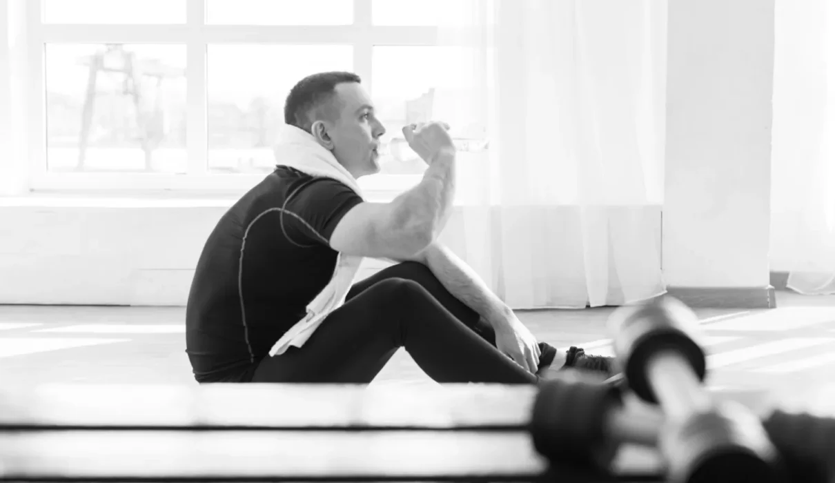 Man sitting alone on a gym bench after a workout under fluorescent lights, looking tired and reflective