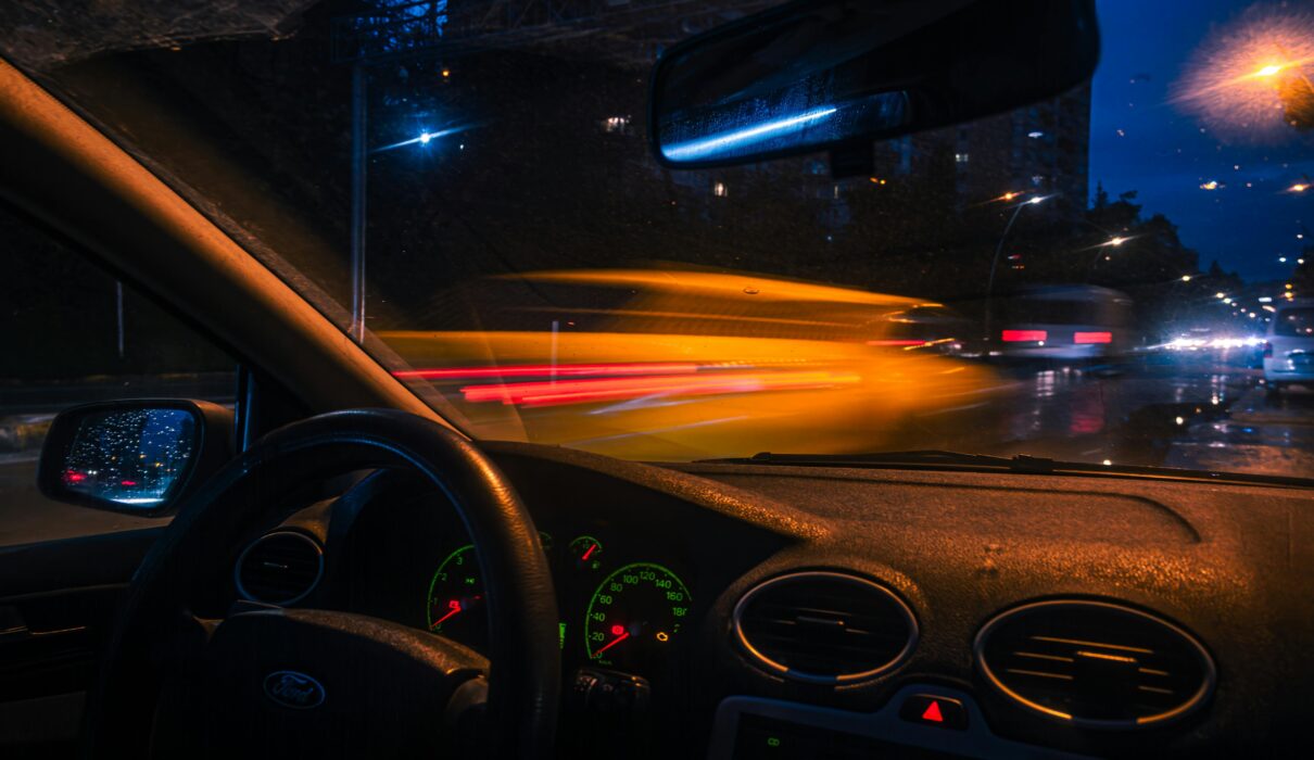 Man sitting alone in a parked car at night outside a gym, staring forward with tired expression and low interior lighting.