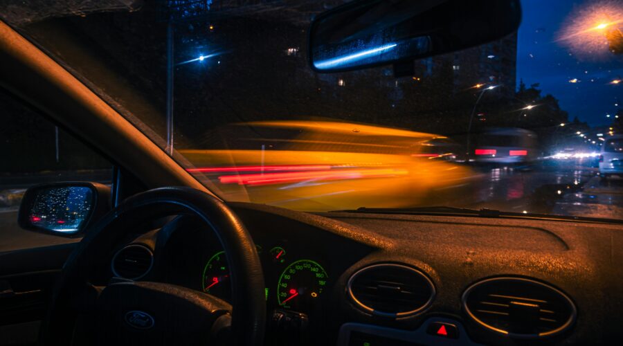 Man sitting alone in a parked car at night outside a gym, staring forward with tired expression and low interior lighting.