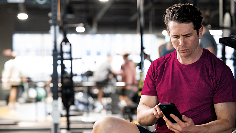 Person standing in gym unsure what exercise to do, checking phone between sets