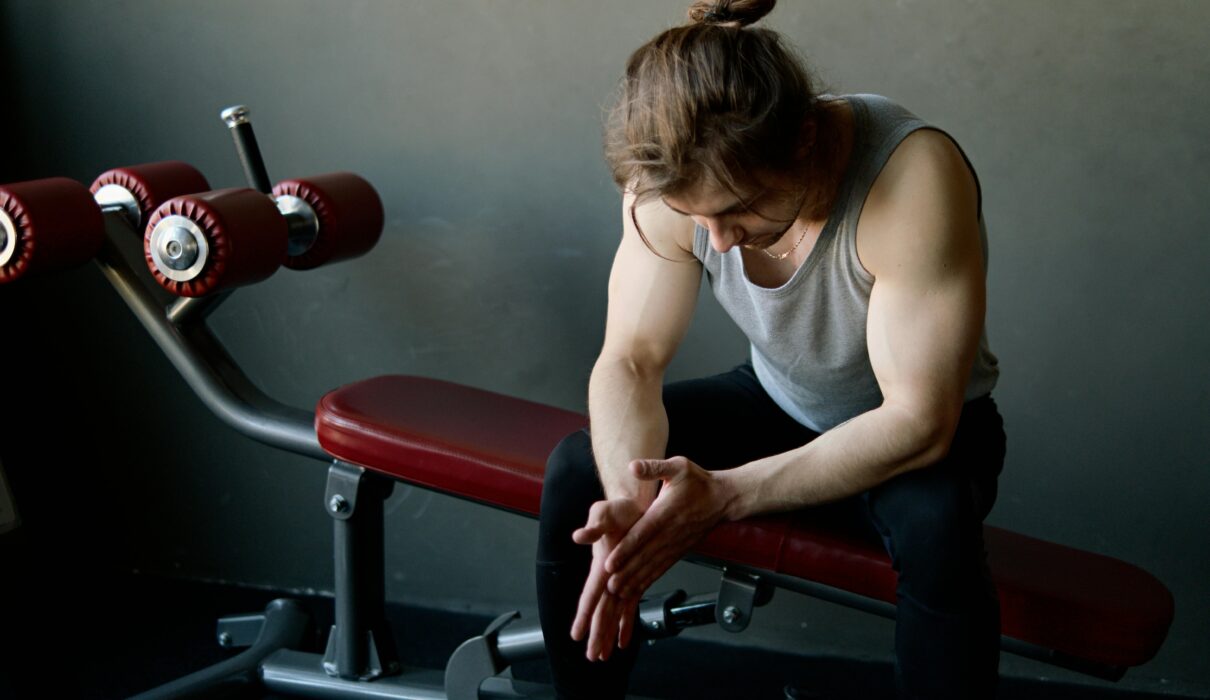 man sitting alone in gym looking tired and thoughtful about fitness consistency